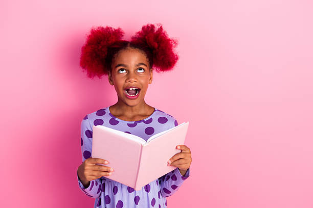 A young girl with vibrant pink hair in a purple polka dot dress holds a pink book and looks up with a playful smile against a pink backdrop.