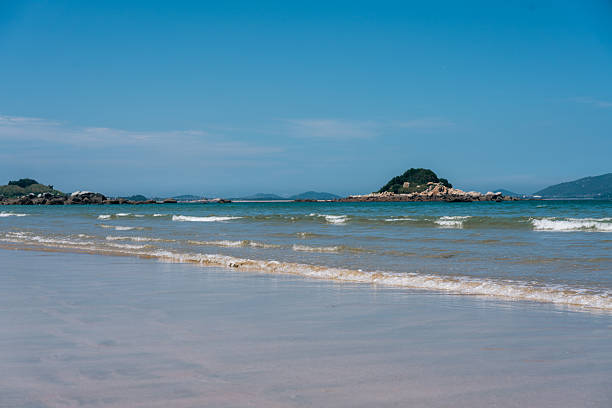 A picturesque coastal scene featuring clear blue sky, gentle waves lapping the sandy shore, and a small island in the distance.