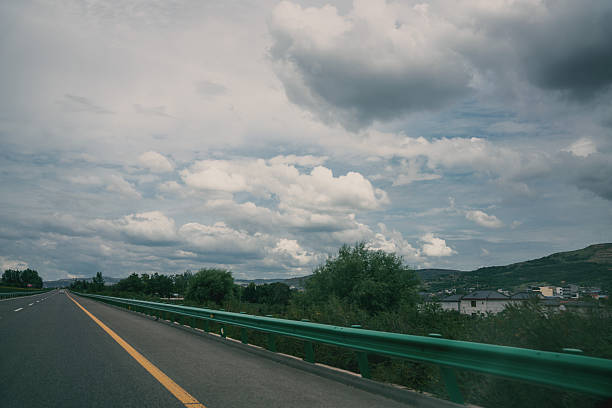 A road with green guardrail under a cloudy sky, showing a natural landscape with trees and distant hills.