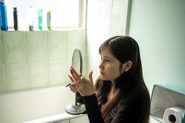 istock Young Woman Looking in a Hand Mirror While Applying Eye Makeup in the Bathroom 2250934235