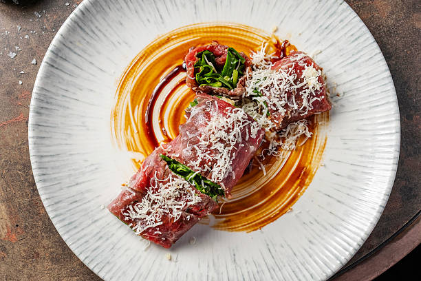 Gourmet beef carpaccio rolls filled with arugula and topped with parmesan shavings, served on a fine dining plate with a painted sauce.