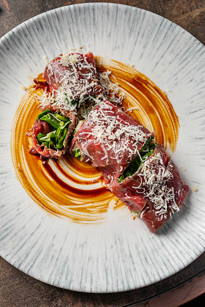 Gourmet beef carpaccio rolls filled with arugula and topped with parmesan shavings, served on a fine dining plate with a painted sauce.
