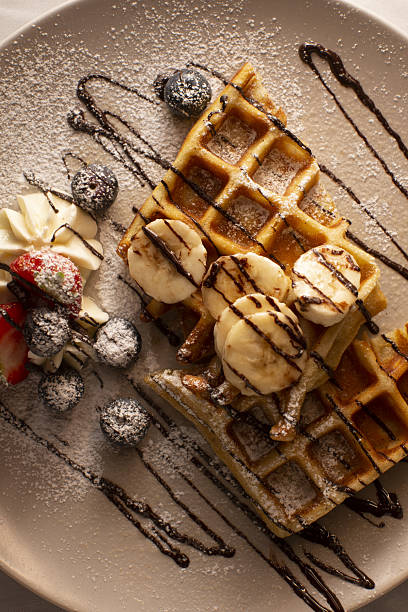 Crispy golden waffles with banana slices and berries, artisan cafe plating with chocolate drizzle, whipped cream, powdered sugar
