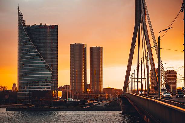RIGA, Latvia – September 01, 2025: Scenic view of an illuminated city skyline and a large architectural bridge crossing water during a beautiful golden hour sunset or dusk.