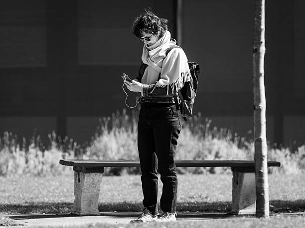 Vienna, Austria - April 7, 2020: A woman stands in a park near a bench and looks at her smartphone, captured in black and white