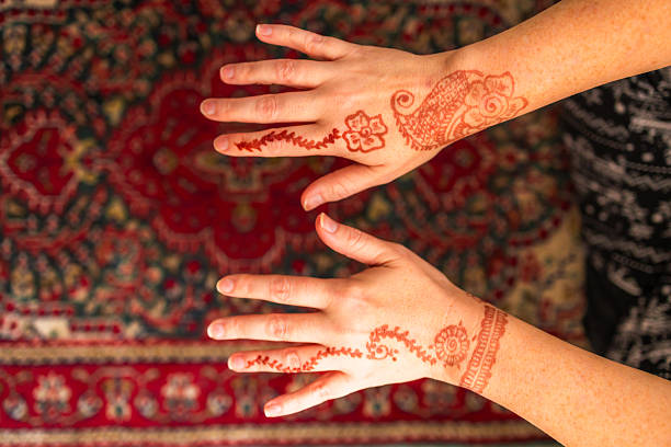 Close up color image depicting an intricate Henna design on a white woman's hands. In the background, defocused, is an Indian rug.
