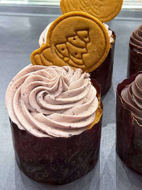 Close-up image bakery display of indulgent desserts, cupcakes in paper cake case on stainless steel tray behind glass panel, chocolate butter icing piped swirls and festive gingerbread Christmas tree cookie decorations, elevated focus on foreground