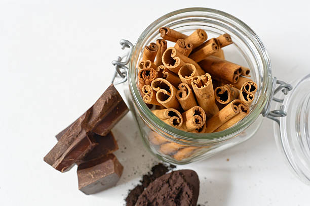 A glass jar filled with cinnamon sticks paired with chocolate pieces and cocoa powder on a spoon, arranged on a clean white background. This simple and warm baking composition highlights natural textures and aromatic ingredients. Copy space on the white surface makes the image suitable for recipe layouts, food branding, and editorial features.