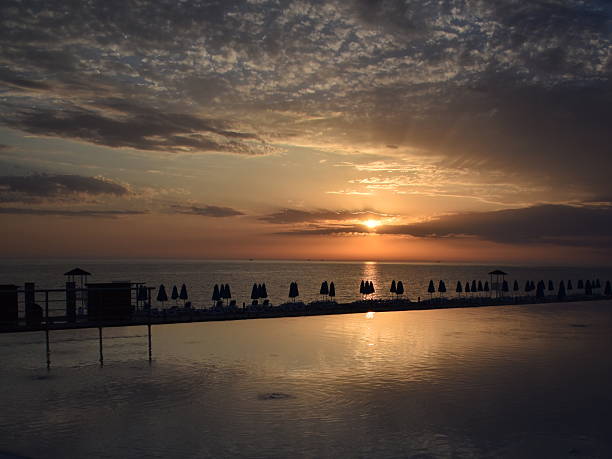 Golden sunset over a seaside beach and hotel swimming pool, with warm sky colors reflecting on calm water. Relaxing coastal evening atmosphere.