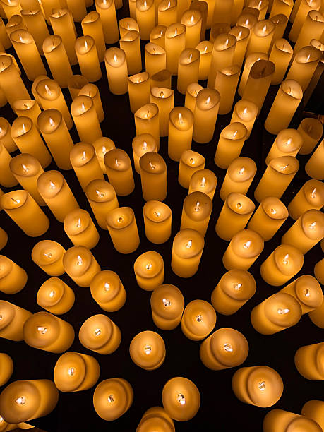 A large collection of flameless LED candles creates a warm, golden glow in a dark setting. The candles vary slightly in shape and height, forming a textured and atmospheric display. The soft, flickering light evokes feelings of peace, remembrance, and serenity, ideal for concepts related to spirituality, memorials, relaxation, and tranquil ambiance.