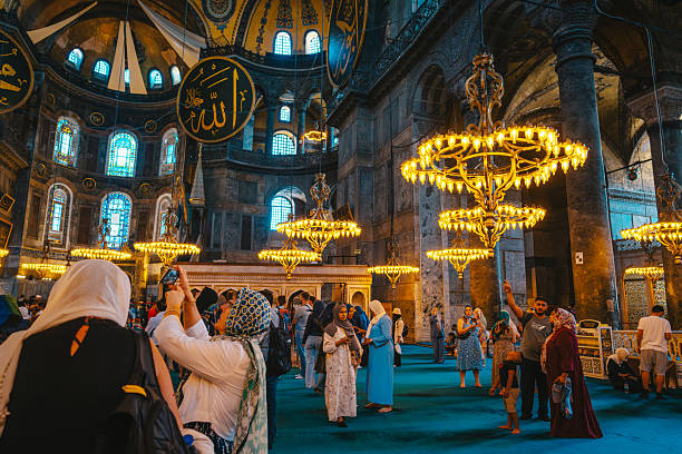 Istanbul, Turkey - June 9, 2022: Iconic Hagia Sophia interior in Istanbul, filled with visitors admiring golden chandeliers, blue carpet, and ornate arches. A vibrant cultural site blending history, architecture, and religious heritage.