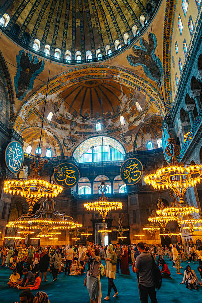 Istanbul, Turkey - June 9, 2022: Dense crowds explore the grand Hagia Sophia, its golden domes and ornate arches inspiring awe. Lamps shimmer over a turquoise carpet as worshippers and tourists share the sacred space.
