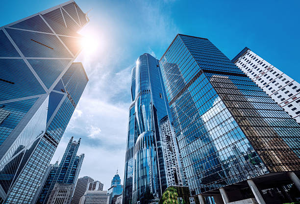 A dramatic view of Hong Kong's glass-clad skyscrapers reaching toward a bright blue sky. The dynamic angle emphasizes urban growth, modern architecture, and vibrant city life.