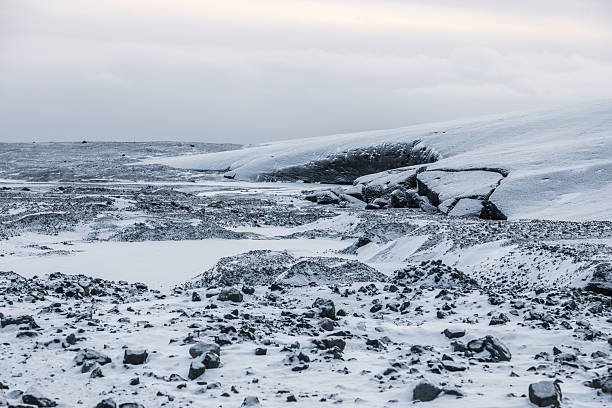 A wide view of the snowy surface of Vatnajokull in Iceland shows the glaciers frozen terrain with exposed ice layers and scattered rocks under a muted winter sky.