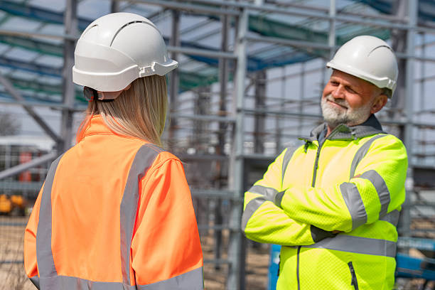 istock Two construction professionals, man and young woman, engaged in conversation at construction site, both wearing safety helmets and high-visibility jackets while reviewing plans on a tablet 2251017815
