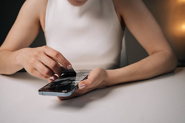 istock Woman prying damaged tempered glass screen protector from smartphone using plastic opening tool, sitting at white table 2251058538