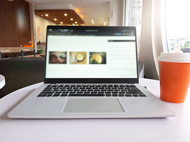 Front view, eye-level shot of an open laptop computer on a white table in a modern, bright coffee shop or cafe. An orange and white disposable takeaway cup sits to the right. The screen displays blurred content, emphasizing the concept of remote work, study, or digital business in a pleasant, public environment