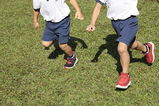 Two children in white shirts and blue shorts run across a sunlit grassy field, casting long shadows as they enjoy outdoor play.