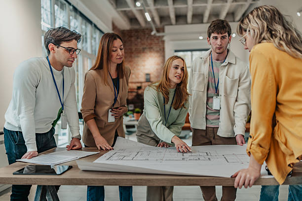 istock Group of young adults discussing office plans in a modern workspace with blueprints on the table 2251077841