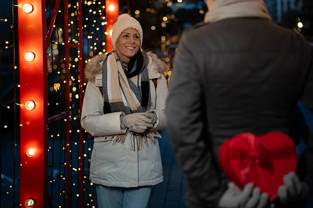 Young man holding a vibrant red heart-shaped gift box behind his back, preparing to surprise his smiling girlfriend during a romantic winter walk in a city lit with festive bokeh lights