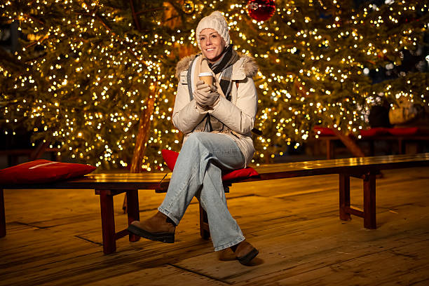 Young adult woman wearing warm winter clothing and holding a coffee cup, sitting on a bench in front of a festive christmas tree decorated with glowing lights