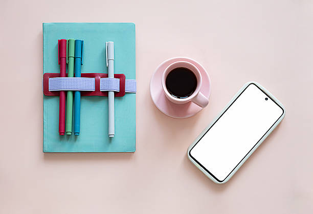 Blue notebook with different colored pens, next to pink coffee cup and smartphone with a white screen for copy space. Desk setup.