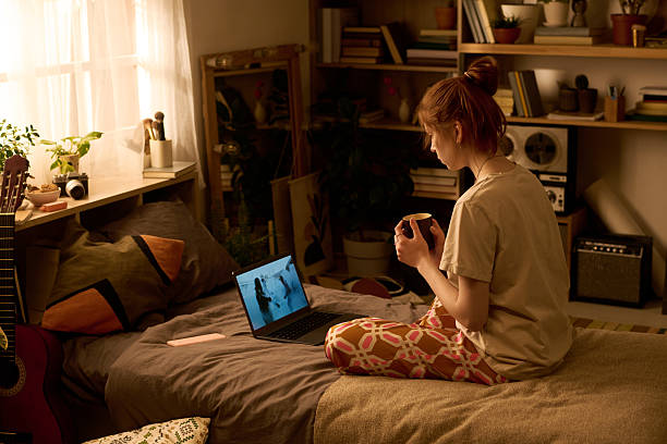 istock Young Woman Sitting on Bed Watching Video on Laptop While Drinking Coffee 2251109856