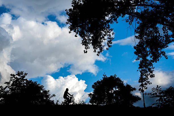 A young Caucasian woman biking across a flat, grassy field with a bicycle, surrounded by a distant tree line and a warm, glowing sky, creating a serene natural setting.
