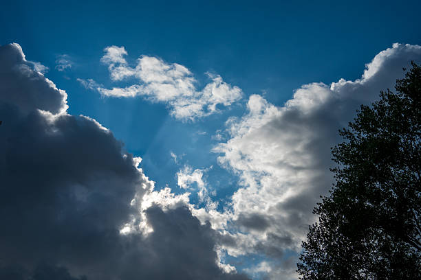 The sky is filled with dark clouds and bright sun rays peaking through, creating an interesting contrast between light and shadow. This cloudy scene captures a serene atmosphere as it features a blue sky above trees below.