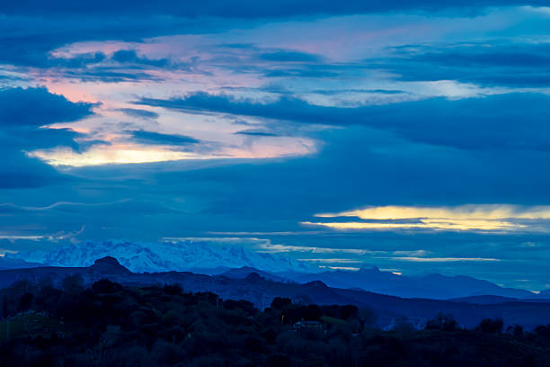 The image features a picturesque mountain scene with a blue sky as the backdrop. In front of a large mountain range, there is a town nestled among the hills. The valley has a mix of greenery and rocky terrain, creating an ideal setting for a quaint village. The scene captures the essence of nature, blending the urban landscape with the grandeur of the mountains. The light shining through the clouds adds to the overall beauty of the image.