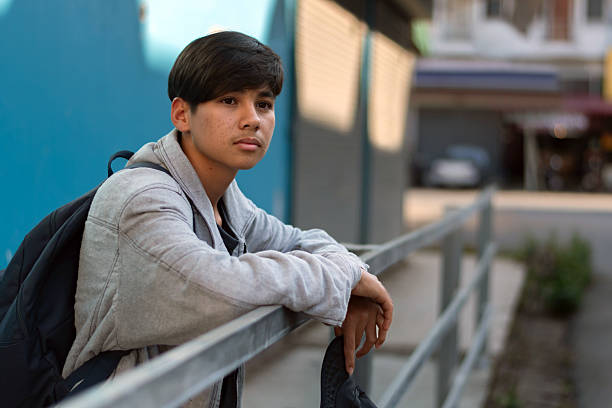 A sad teen in a grey long-sleeve denim jacket, carrying a backpack, standing and waiting for someone at a gloomy bus station, escaping family pressure for a new life and broken home concepts.
