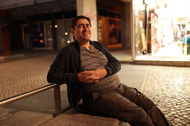Mature man sitting on a stone bench, smiling contentedly in a vibrant urban street at night