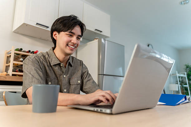istock Asian attractive man freelancer using laptop device working in house. Handsome male student chatting and typing on computer notebook to communicate online with technology in cozy kitchen at home. 2251137616