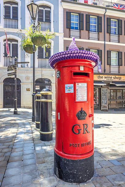 Gibraltar, United Kingdom - September 10, 2024: Victorian post box of standard UK design. A red postbox adorned with a knitted hat sits on a cobblestone street, in front of historic buildings.