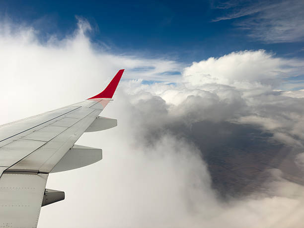 A serene and dynamic view from a passenger window, capturing the metal wing and red winglet of an airplane soaring high above a sprawling blanket of thick, white cumulus clouds. The bright sunshine highlights the texture of the clouds, creating a sense of depth and peaceful isolation, contrasting with the deep blue sky above. This image evokes feelings of travel, freedom, adventure, and the journey itself, making it perfect for themes related to flight, exploration, and perspective.