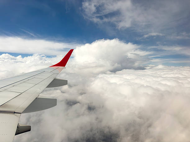 A serene and dynamic view from a passenger window, capturing the metal wing and red winglet of an airplane soaring high above a sprawling blanket of thick, white cumulus clouds. The bright sunshine highlights the texture of the clouds, creating a sense of depth and peaceful isolation, contrasting with the deep blue sky above. This image evokes feelings of travel, freedom, adventure, and the journey itself, making it perfect for themes related to flight, exploration, and perspective.