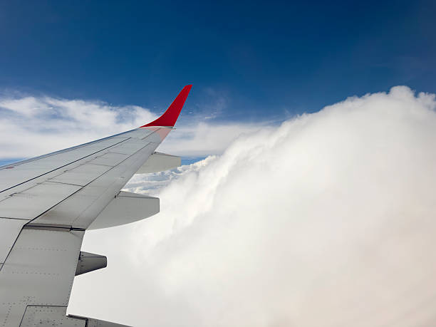 A serene and dynamic view from a passenger window, capturing the metal wing and red winglet of an airplane soaring high above a sprawling blanket of thick, white cumulus clouds. The bright sunshine highlights the texture of the clouds, creating a sense of depth and peaceful isolation, contrasting with the deep blue sky above. This image evokes feelings of travel, freedom, adventure, and the journey itself, making it perfect for themes related to flight, exploration, and perspective.