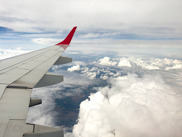 A serene and dynamic view from a passenger window, capturing the metal wing and red winglet of an airplane soaring high above a sprawling blanket of thick, white cumulus clouds. The bright sunshine highlights the texture of the clouds, creating a sense of depth and peaceful isolation, contrasting with the deep blue sky above. This image evokes feelings of travel, freedom, adventure, and the journey itself, making it perfect for themes related to flight, exploration, and perspective.