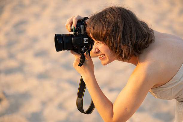 istock Young woman smiling and focused while holding a dslr camera, capturing golden-hour beach scenes on warm sand, backlit, relaxed and passionate about photography hobby 2251146644