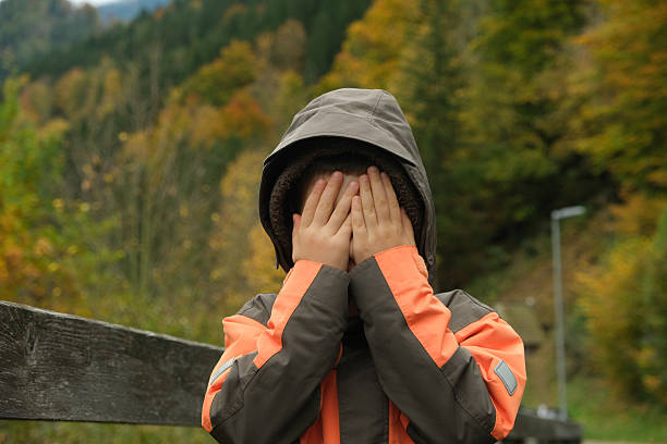 istock Shy Little Boy Covering His Face With His Hands While Walking Through an Autumn Park 2251147366