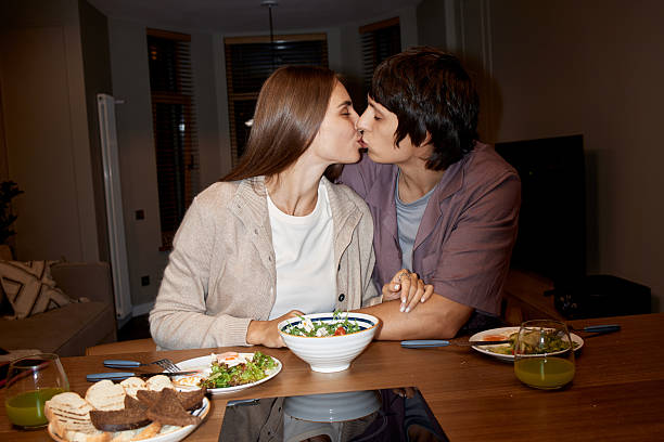 Young adult Caucasian woman and young adult Caucasian man kissing while sitting at dining table, holding hands, sharing meal with salad, bread, and green juice visible on table