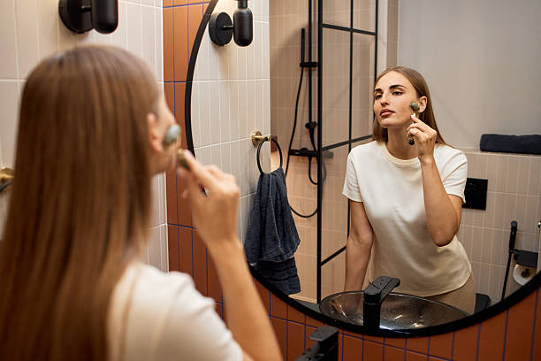 istock Young Woman Using Facial Roller While Looking into Bathroom Mirror 2251155895