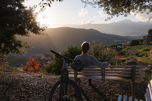 Young male mountain biker  sits on bench above hills at sunset and looks out, Finale Ligure, Liguria