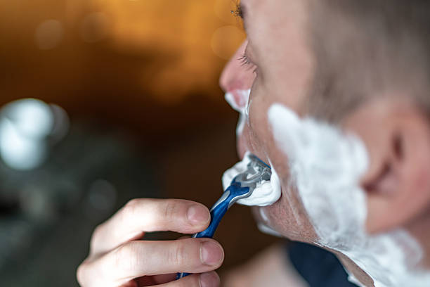Man removing shaving foam with a disposable razor during a morning grooming routine in the bathroom, close-up on face and hand showing clean-shaven, fresh personal care ritual