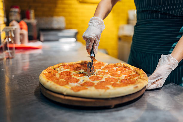 Female worker slicing pizza into portions using a pizza wheel. Fast food service, food preparation detail, and commercial kitchen environment