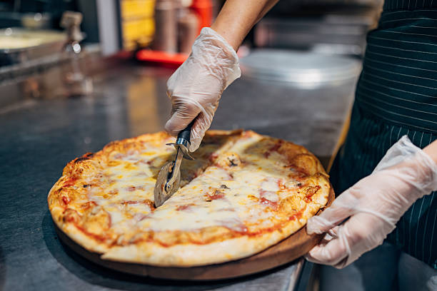 Unrecognizable female worker cutting pizza into portions for customer service. Quick service restaurant, takeaway food preparation, and commercial kitchen environment