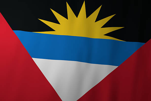 The image shows the national flag of Antigua and Barbuda, featuring a rising yellow sun set against a black background. Beneath it are blue, white, and red sections symbolizing the sea, sand, and the strength of the people.