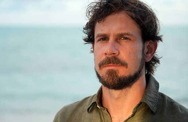 Man with a beard posing for a head shot on the beach wearing casual clothes