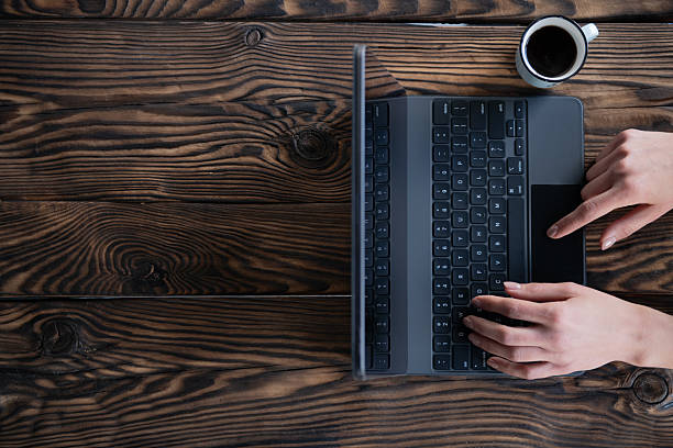 Minimal lifestyle scene with female hands typing on a laptop next to a cup of coffee on a rustic wooden table. A clean work-from-home setup conveying productivity, remote work and modern digital lifestyle.