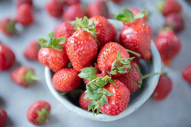 Top-down macro of ripe strawberries in a handmade ceramic bowl surrounded by scattered berries. Natural soft lighting and shallow depth of field ideal for food styling, nutrition themes, summer produce and organic lifestyle concepts.